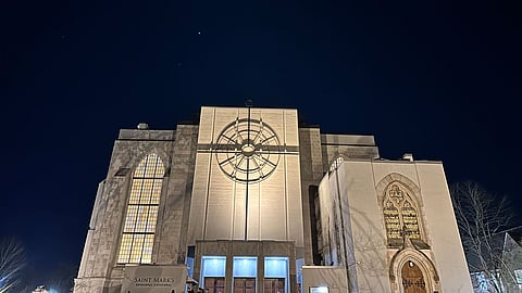 A photo of Seattle’s St Marks Cathedral at night.  The  cathedral has its lights on, illuminating it’s glass window features.  A large cross featured on the front of the building is also illuminated.