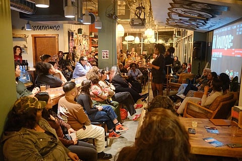 A woman speaks at a microphone in a warmly lit café-style venue, addressing a diverse audience seated on chairs and couches. A large screen on the right displays event branding with the words “lit lounge.”