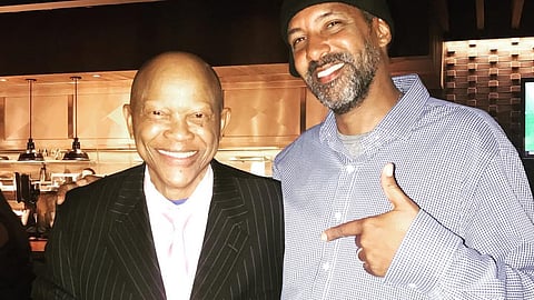 Two smiling men posing together in a warmly lit restaurant. The man on the left wears a pinstriped suit with a pink tie and points toward the man on the right, who wears a checkered shirt and black beanie, also pointing back at the first man.