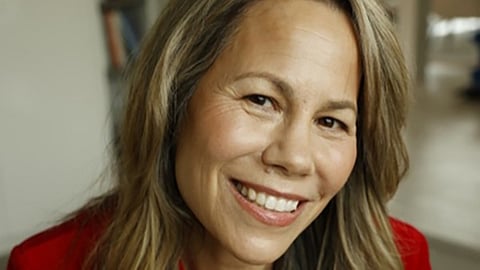Smiling woman with long, light brown hair wearing a red blouse, posing indoors with a blurred background.