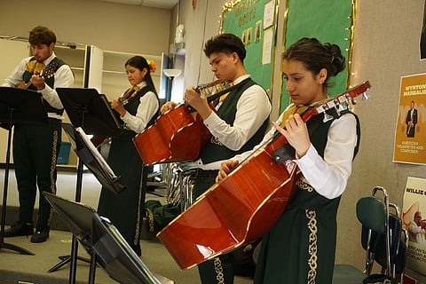 A group of mariachi students in green and gold uniforms play stringed instruments in a classroom, focused on their music.
