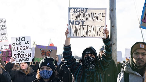Protesters gathered outdoors holding signs advocating for immigrant rights. The central sign reads “Fight Ignorance Not Immigrants.” Other visible signs include “No One Is Illegal on Stolen Land” and a butterfly illustration.