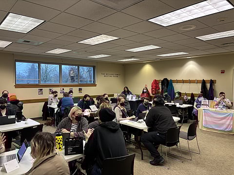 Volunteers help people with paperwork for name and gender-marker changes at the Gender Justice League clinic at the Columbia City library on Feb. 23. People are sitting around tables in chairs and speaking to one another, many of them masked.