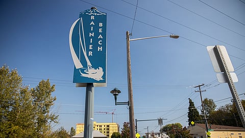 Street sign reading “Rainier Beach” with a sailboat graphic and Mount Rainier silhouette stands near a sidewalk under a clear blue sky. In the background are residential houses, a construction site, and utility poles lining the quiet urban street.