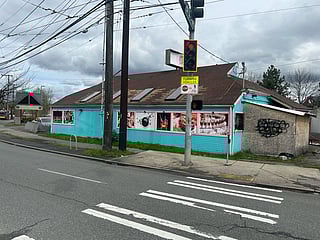A turquoise and pink building on a street corner displays food and drink posters in its windows, with one boarded-up and graffiti-covered section on the right. A red traffic light and crosswalk signal are visible at the intersection in front.