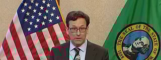 The Washington State governor, a white man wearing glasses and a suit, speaks at a podium with a mic. Washington State and U.S. flags are draped behind him.