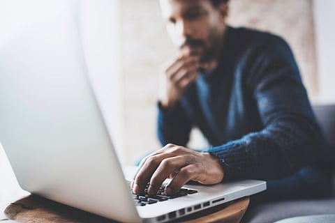 A man is reading on a laptop, his hand resting on the keyboard, his other hand touching his bearded chin.
