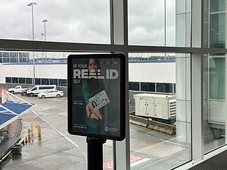 Poster in an airport terminal promoting REAL ID compliance, showing a woman holding out a REAL ID card with the message “Be your REAL ID self” and a reminder to be ready by May 7, 2025. Rainy tarmac and airport vehicles are visible through the large windows behind it.