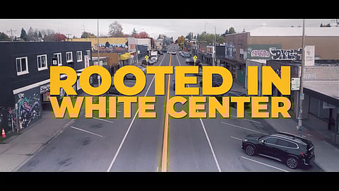 Aerial view of a street in the White Center neighborhood lined with small businesses and street art, overlaid with bold yellow text that reads “ROOTED IN WHITE CENTER.”