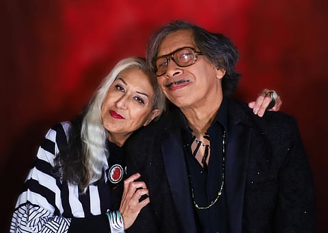 Portrait of a smiling couple posed in front of a rich red background. The woman rests her head on the man's shoulder, wearing a black and white patterned outfit with silver and red jewelry, while the man wears glasses and a black suit with beaded necklaces.