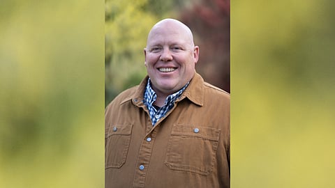 A headshot of Jamie Fackler shows him wearing layered button-up shirts and smiling at the camera against a backdrop of blurred foliage.
