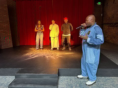 A group of four individuals stands on a stage with red curtains in the background. One person in the foreground, dressed in a blue jumpsuit, speaks into a microphone while addressing the group.