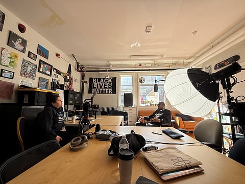Behind-the-scenes view of a video interview in progress inside a creative workspace, featuring a "Black Lives Matter" banner, studio lights, and camera equipment, with two people engaged in conversation across a large table.