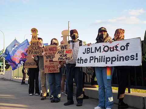 Protesters holding signs and cardboard cutouts shaped like chess pawns that read "We are not your pawns" and "JBLM out of the Philippines!" while standing on a sidewalk, some wearing masks and holding a large blue flag.