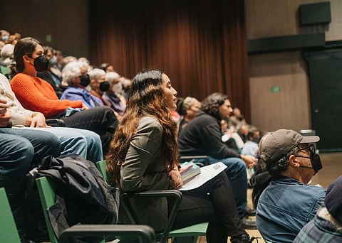 Audience members seated in rows inside a theater or auditorium, attentively watching an event; several people are wearing face masks, and one woman in the foreground holds a booklet and pen.