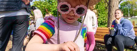 A young child wearing a straw cowboy hat with a pink star, rainbow-striped sleeves, and flower-shaped sunglasses looks closely at a ladybug crawling on their hand during an outdoor event.