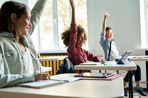 Three students sit at desks in a bright classroom, smiling and raising their hands eagerly to participate; notebooks and a tablet are visible on their desks.