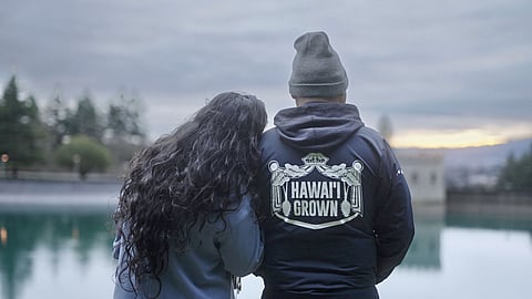 Two people stand close together facing a calm body of water at dusk, one resting their head on the other’s shoulder; the back of the person’s jacket reads “HAWAI‘I GROWN” with graphic elements around the text.