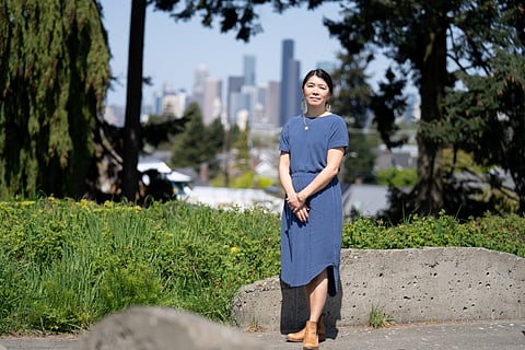 An Asian woman in a blue dress and brown boots stands near greenery. Conifer trees and the Seattle skyline are in the background.