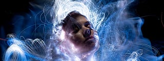 A lightpainting portrait around a person's face at long exposure; the person has their eyes closed and their face tilted upward, and the lightpainting around them gives the image an ethereal look.