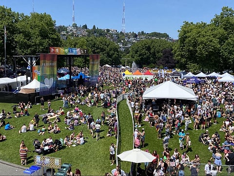 Large crowd gathered outdoors for PrideFest at a park, with colorful tents, banners, and a rainbow-themed stage.