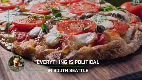 Close-up of a rustic-style pizza topped with anchovies, fresh tomato slices, melted mozzarella, basil leaves, and capers on a wooden board. Overlay text reads “EVERYTHING IS POLITICAL IN SOUTH SEATTLE” with a small inset image of a man in the lower-left corner.