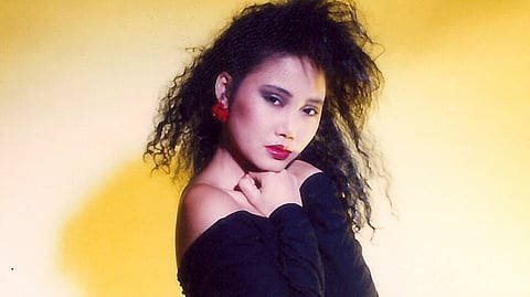 Portrait of a Vietnamese woman in dramatic 1980s-style fashion, wearing a black off-the-shoulder top, red flower earrings, and bold makeup. She has voluminous, crimped hair and poses against a warm yellow backdrop.