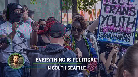 A Black woman reaches her hand out toward a white man whose back is turned to the camera. Protesters and counterprotesters with signs stand around them.