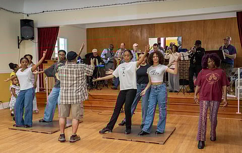 Group of young tap dancers performs choreographed moves on wooden platforms in front of a live jazz band.