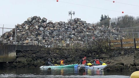 Kayakers paddle near a large scrap metal heap at an industrial waterfront site, with fencing, power lines, and pollution concerns in the background.