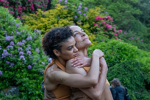 Two contemporary dancers embrace in a lush botanical garden surrounded by blooming rhododendrons.