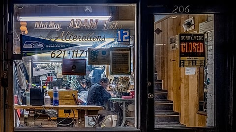 Nighttime view of Adam Tailor Alterations in Seattle’s Pioneer Square, showing tailors working inside a small garment shop filled with sewing tools, clothing racks, and vintage signage.