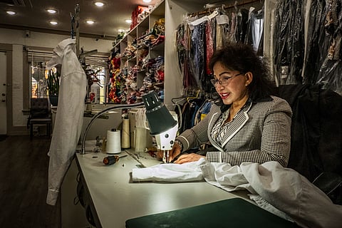 Vietnamese woman tailor sewing white shirt in fabric shop; woman at sewing machine with thread spools, garments, and fabric rolls in background.