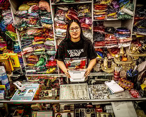 Young Vietnamese woman at fabric and jewelry counter in tailor shop, standing in front of shelves stacked with colorful folded textiles and traditional Vietnamese accessories.