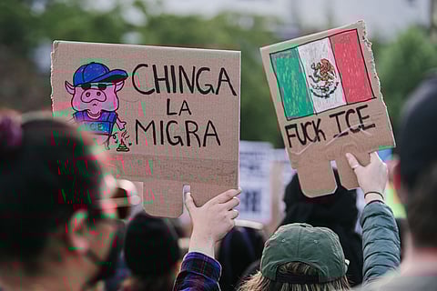 Anti-ICE protest in Seattle with demonstrators holding bilingual signs criticizing U.S. immigration enforcement, including cartoon imagery and the Mexican flag.