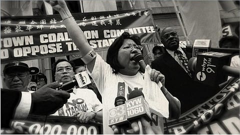 A still from "Taking Root: Southeast Asian Stories of Resettlement in Philadelphia" shows a woman speaking into a microphone while other people hold microphones around her; her arm is raised into the air and she's speaking passionately. Other people holding signs are standing behind her.