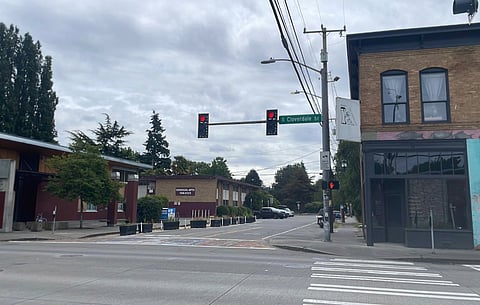An empty street corner. Street lights are red. A tan brick two-story building is in the foreground to the right. A more modern brick red building is to the left and tan two-story apartments are in the distance.