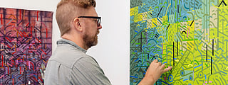 Artist painting detailed geometric abstract artwork in studio, vibrant green and blue patterns, with art supplies and another colorful piece on wall at J Rinehart Gallery.