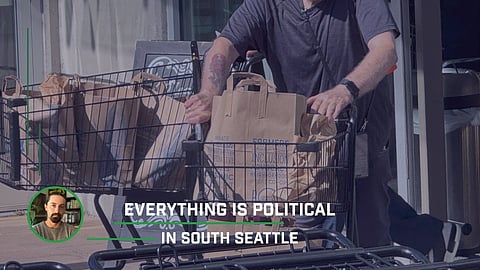 Person pushing grocery carts filled with brown paper bags at PCC Community Markets in Columbia City, with “Everything is Political in South Seattle” text overlay.
