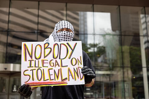 Protester in face covering holds sign saying “Nobody illegal on stolen land” during demonstration in front of a glass building in downtown Seattle.