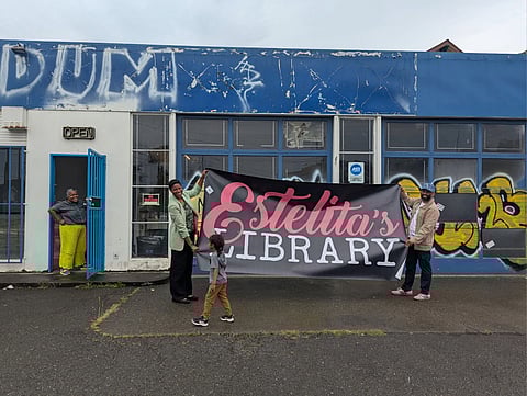 Estell Williams and Edwin Lindo hold a sign that reads "Estelita's Library" between them against the windows of the former auto shop; a child stands in front of the sign facing Estell.
