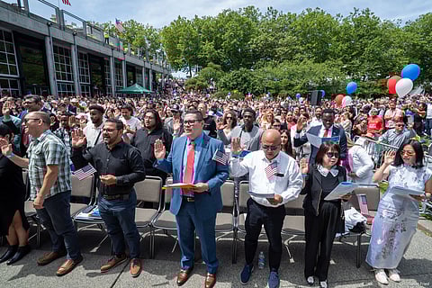 New U.S. citizens taking oath at outdoor naturalization ceremony, holding American flags, with large diverse crowd and patriotic decorations.