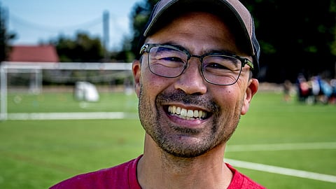 Eddie Lin, Seattle District 2 candidate, smiling outdoors on a soccer field while wearing glasses, a red shirt, and a cap with a rainbow pride patch.