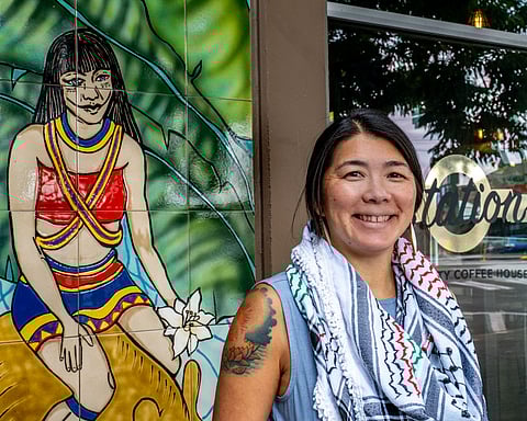 Jeanie Chunn standing outside The Station coffeehouse in Seattle, smiling beside a vibrant mural of an Indigenous woman. She wears a keffiyeh scarf and a sleeveless top.