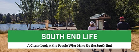 Three people walk along a paved path at Seattle's Seward Park on a sunny day, with Lake Washington and trees in the background. A park map kiosk stands beside the trail, and dry summer grass lines the walkway.