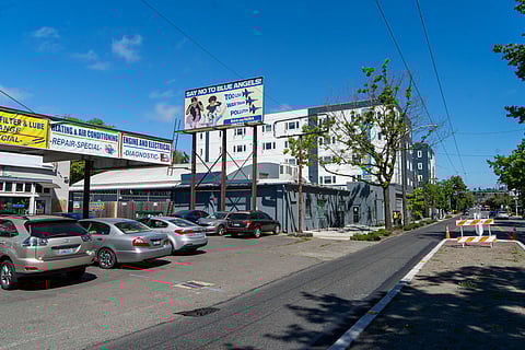 A Seattle street scene shows a large billboard reading “Say No to Blue Angels” above a parking lot with several cars. Nearby banners advertise auto repair services, and multi-story apartment buildings line the street.