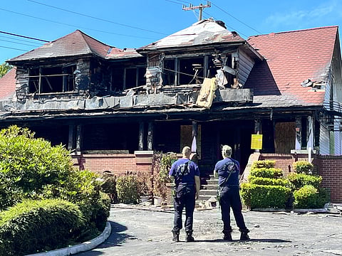 Two firefighters stand in front of a heavily fire-damaged building with a red roof and boarded-up windows at Columbia Funeral Home. The charred structure shows extensive fire destruction on the upper floor and roof.