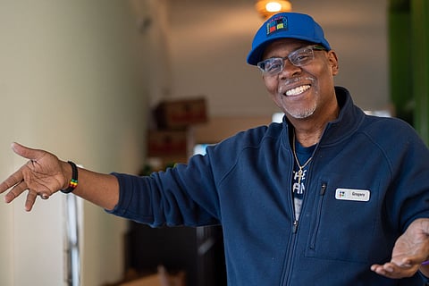 A Black man wearing a blue baseball cap and blue fleece jacket greets the viewer with open arms.