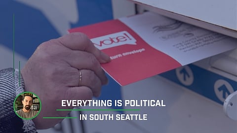 Close-up of a voter inserting a red "Vote!" ballot envelope into an official ballot drop box, with a small inset photo of a man in the lower-left corner and the text "Everything is Political in South Seattle" overlaid at the bottom.