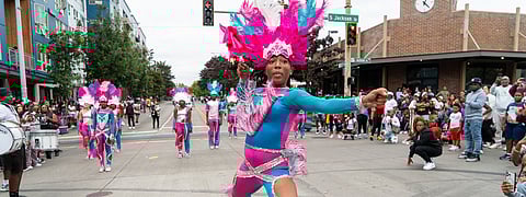 Performer in a vibrant feathered headdress and pink-blue fringe costume leads a street drill team routine during the 2025 Umoja Fest parade at S. Jackson Street in Seattle, with cheering spectators and a drumline in the background.